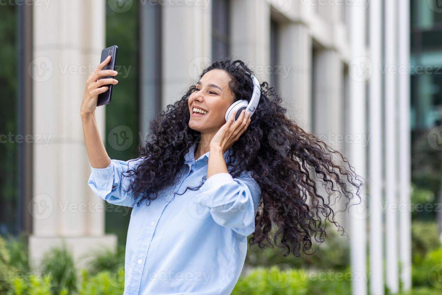 een vrolijk jong vrouw met gekruld haar- en hoofdtelefoons is glimlachen terwijl nemen een selfie buitenshuis met haar smartphone. ze straalt uit geluk en vertrouwen in een gewoontjes instelling. foto
