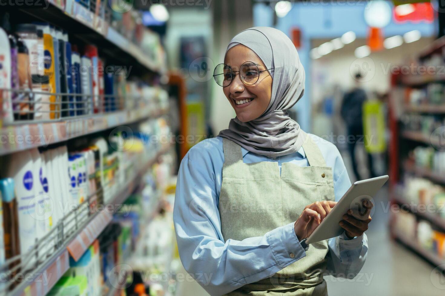 een vrouw cheques producten en goederen in een supermarkt, een moslim vrouw in een hijab toepassingen een tablet in de buurt een plank met goederen, een arbeider in een schort glimlacht Bij werk foto