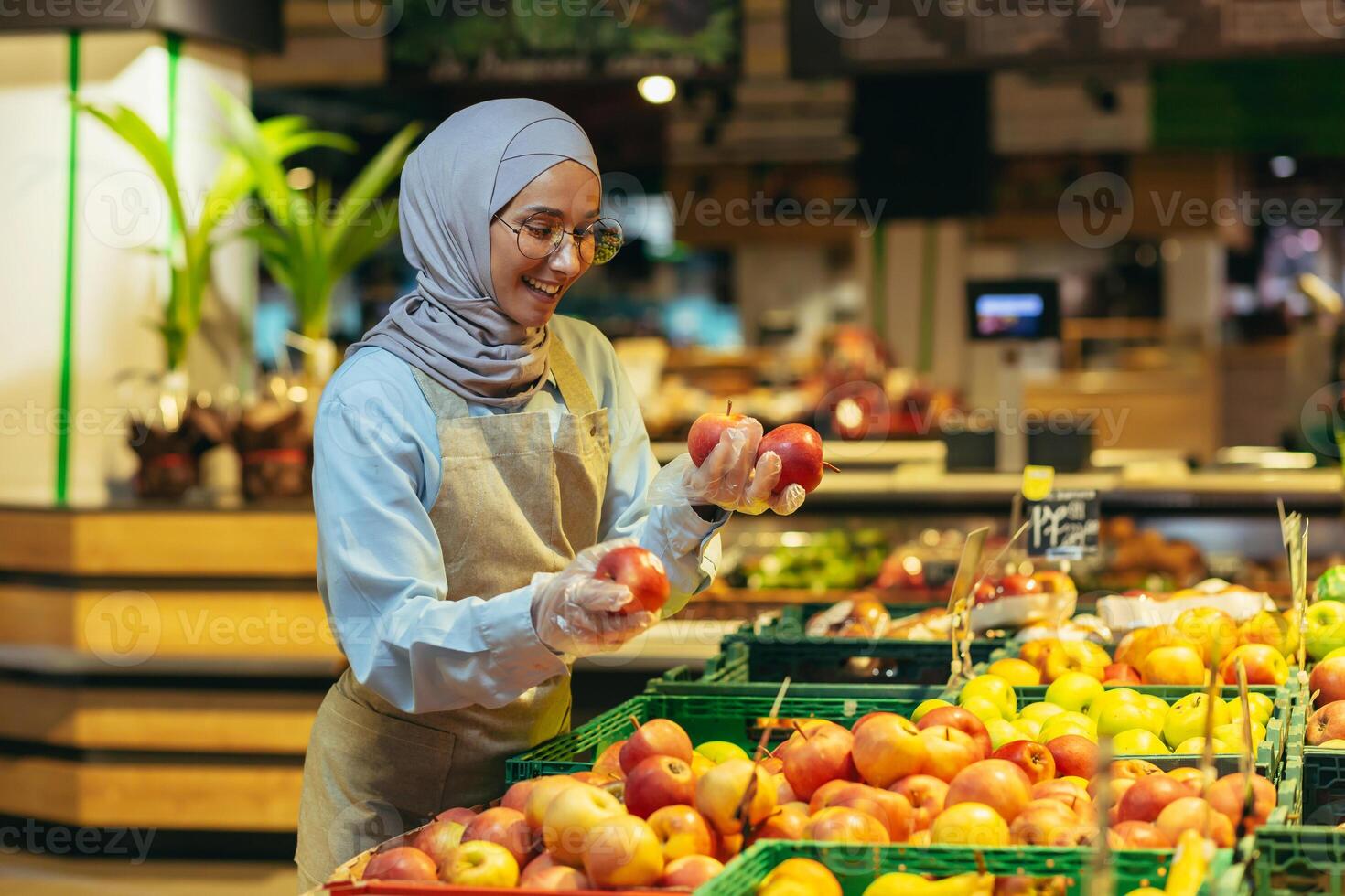 vrouw verkoper in hijab browsen en controle appels in supermarkt, vrouw in schort glimlachen Bij werk in op te slaan in fruit en groente afdeling foto