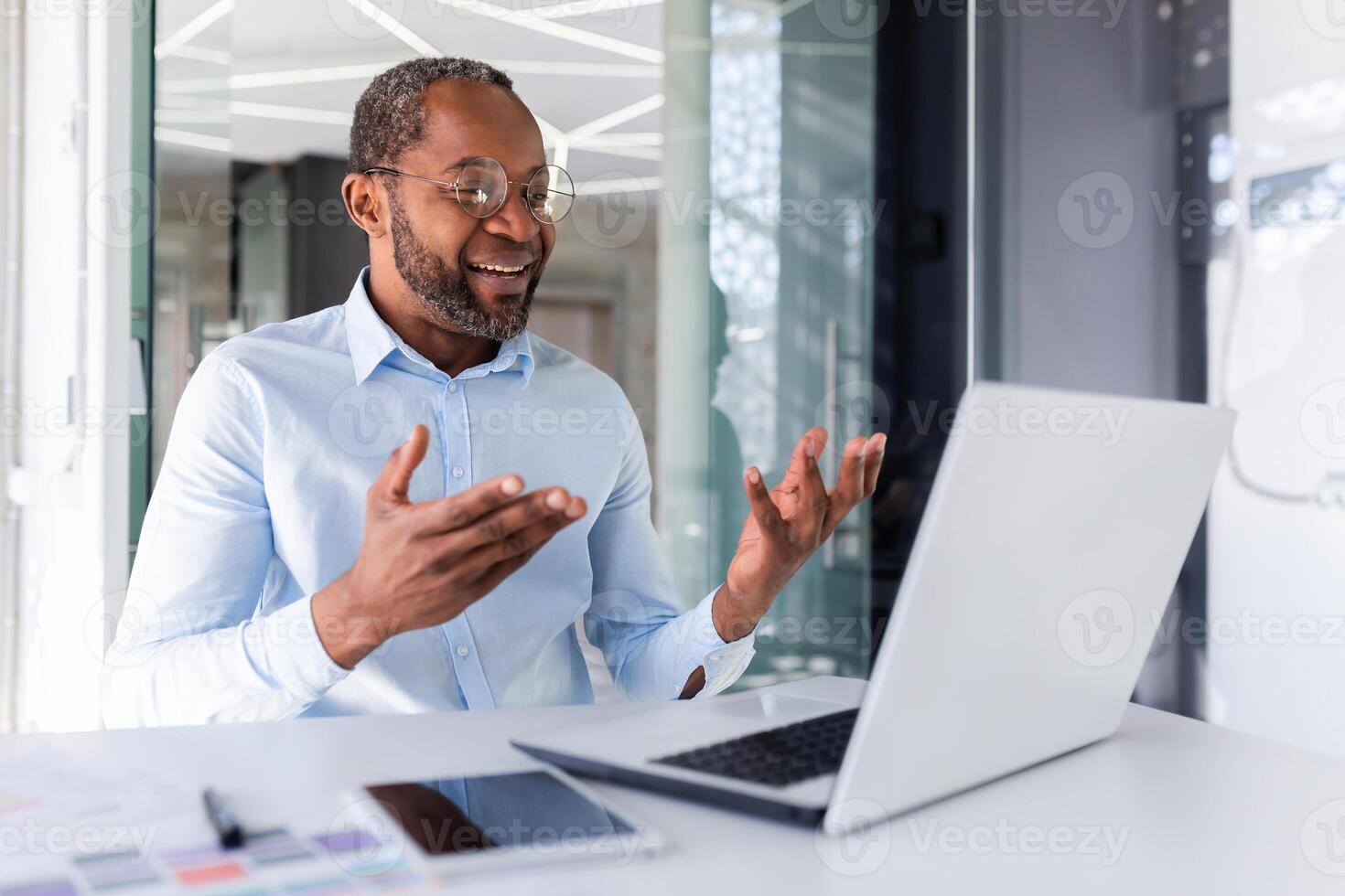 volwassen Afrikaanse Amerikaans baas werken binnen kantoor met laptop van een afstand, zakenman in overhemd Aan online vergadering met collega's, Mens pratend Aan telefoontje gebruik makend van laptop terwijl zittend in shirt. foto