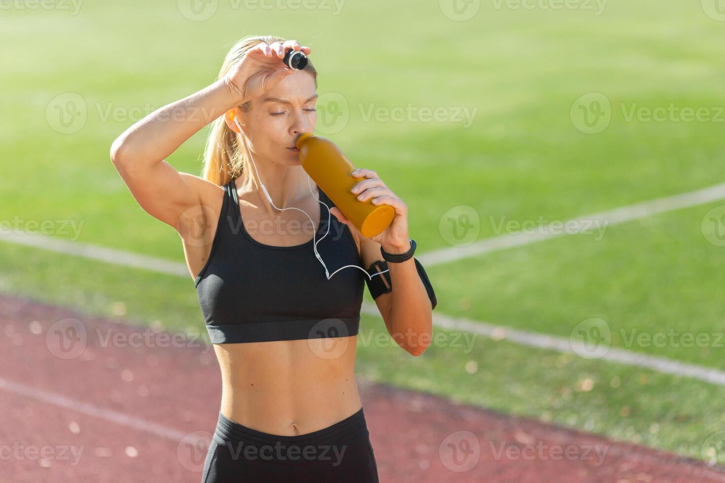 een fit jong vrouw met oortelefoons is nemen een breken naar hydrateren met een water fles Aan een zonnig atletisch spoor. foto