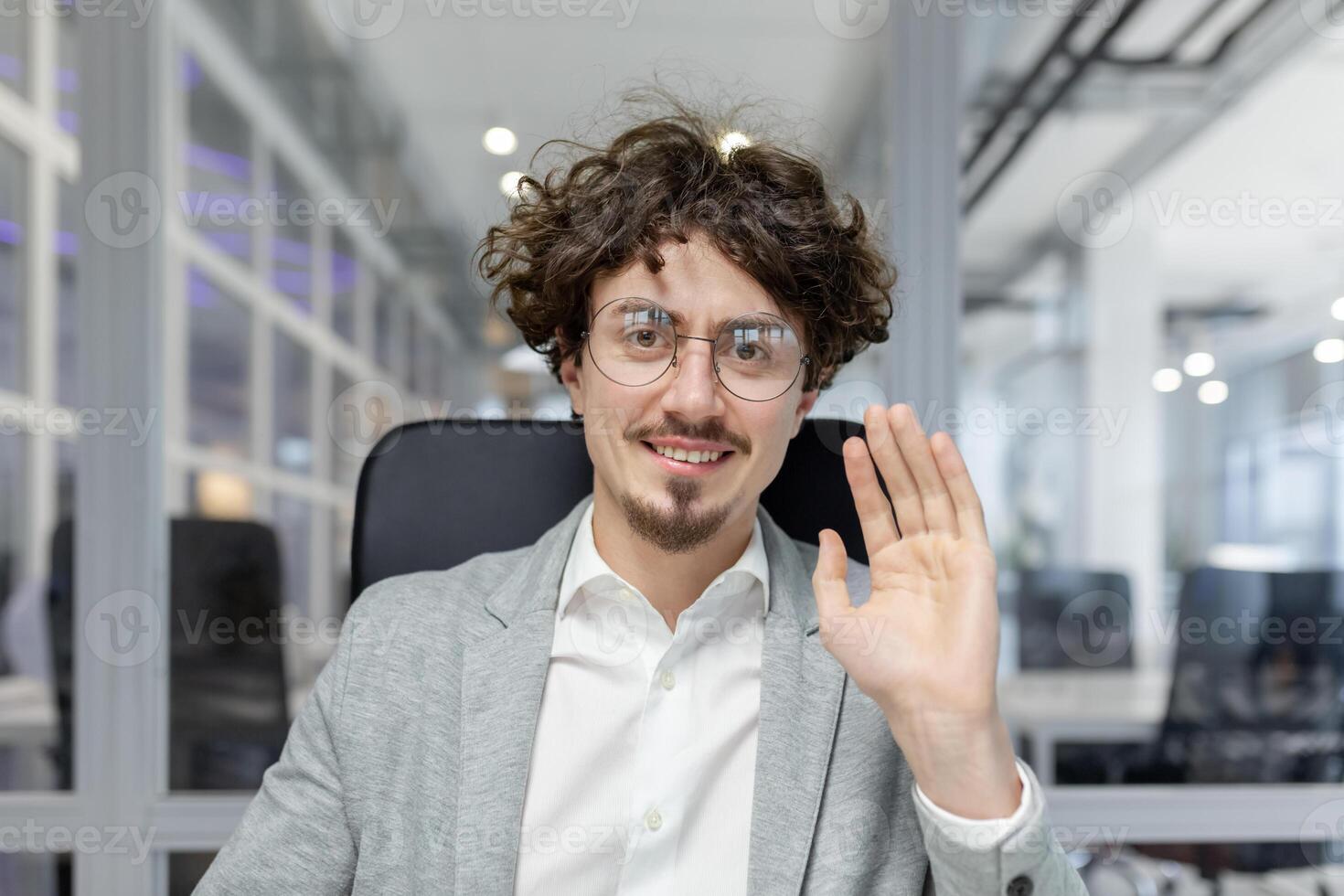 een professioneel jong volwassen zakenman in slim kleding golven Hallo, tonen positiviteit en vertrouwen temidden van een zakelijke instelling. foto