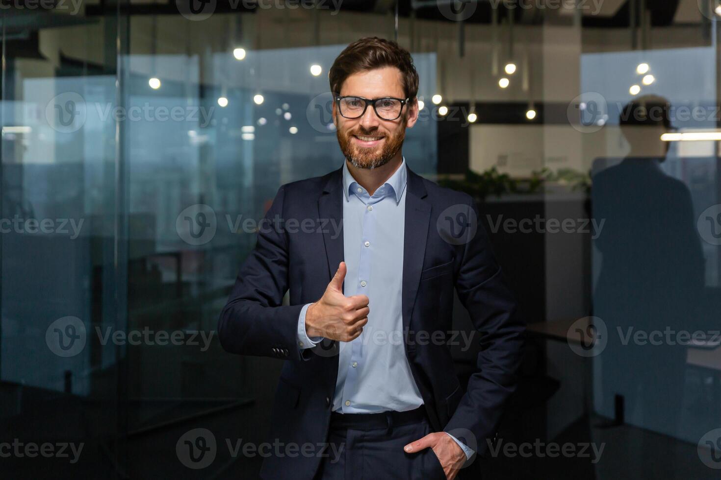 portret van geslaagd volwassen zakenman baas, manager in bedrijf pak bril en baard op zoek Bij camera en glimlachen staand in de buurt venster, tonen duimen omhoog, teken van succes en bereiken doelen. foto