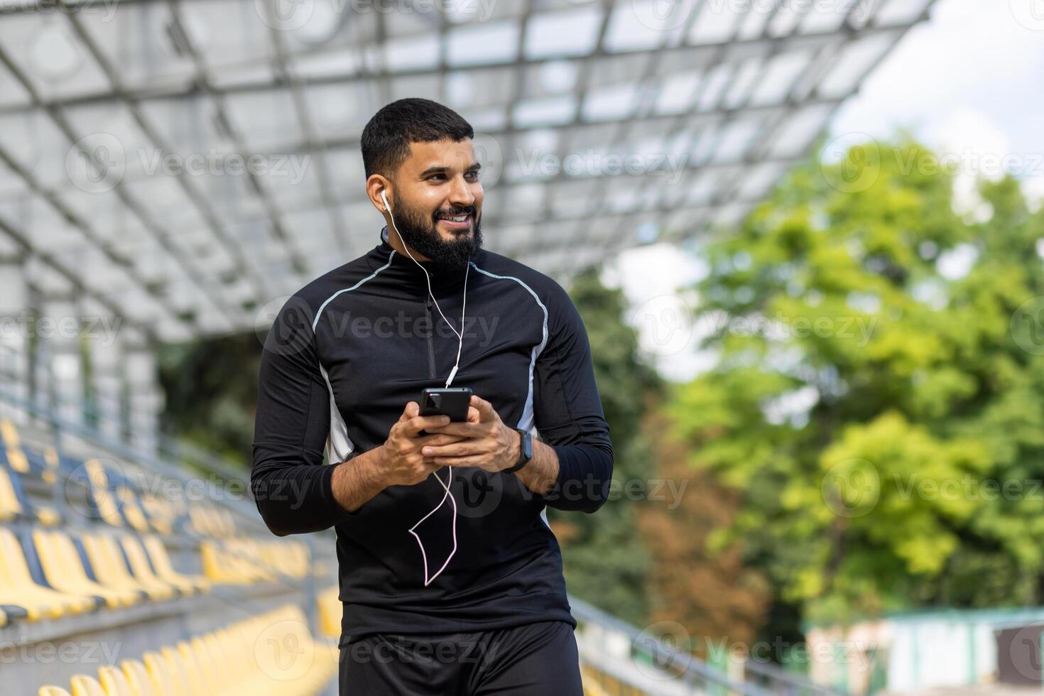 een lachend, gebaard Mens vervelend sportkleding en oortelefoons houdt een smartphone terwijl staand Bij een buitenshuis stadion. foto