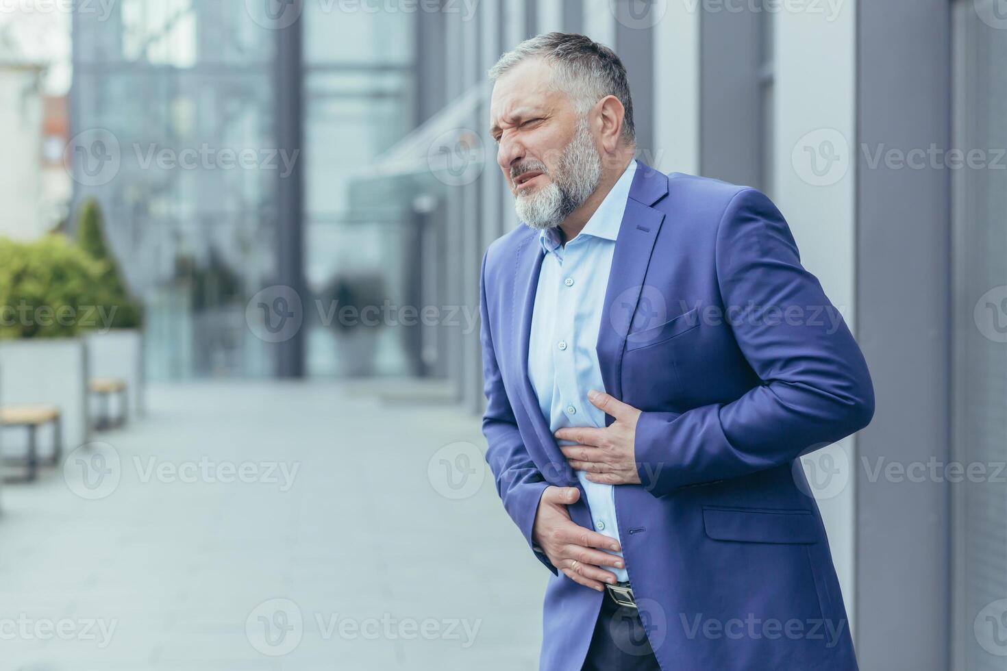 senior Mens, zakenman, kantoor arbeider in een bedrijf pak staat in de buurt de kantoor centrum Aan de straat en houdt zijn maag met zijn handen. hij voelt erge, ernstige pijn in zijn maag, hij behoeften helpen. foto