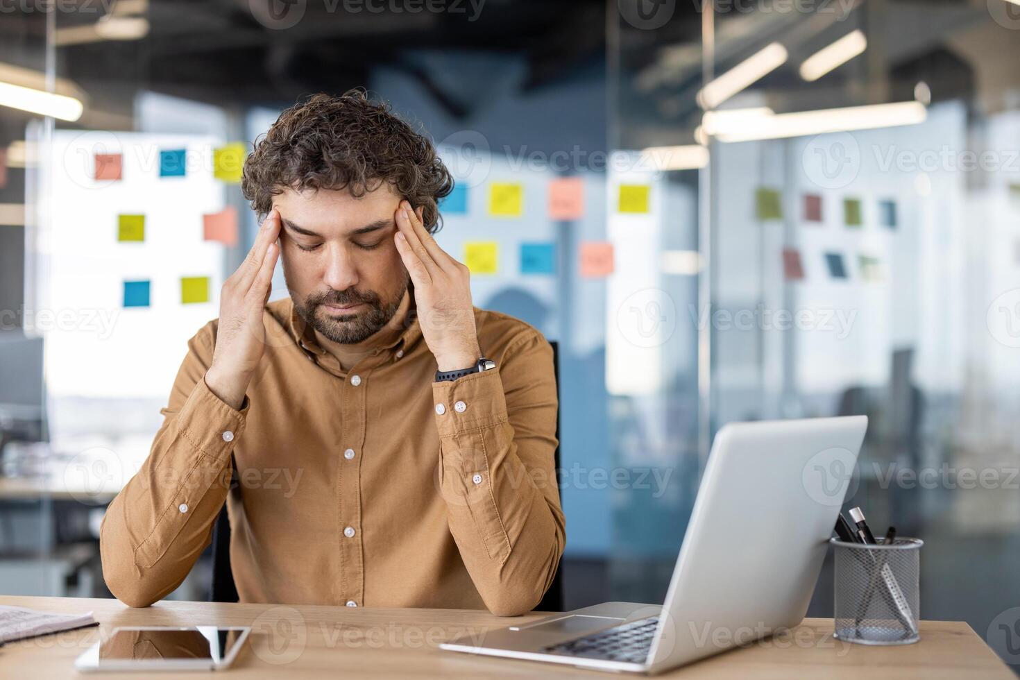 een mannetje professioneel in een bruin overhemd voelt pijn, Holding zijn hoofd in nood terwijl zittend Bij zijn kantoor bureau, wijzend op spanning of migraine. foto