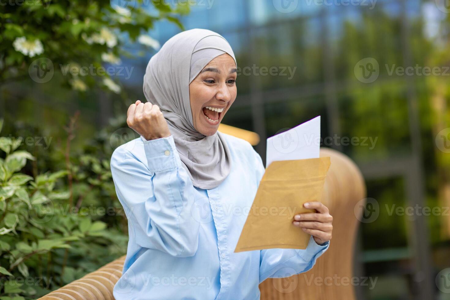 blij jong vrouw vervelend een blauw hijab buitenshuis, vieren succes met een envelop in hand, uitdrukken geluk en positiviteit in een stedelijk instelling. foto