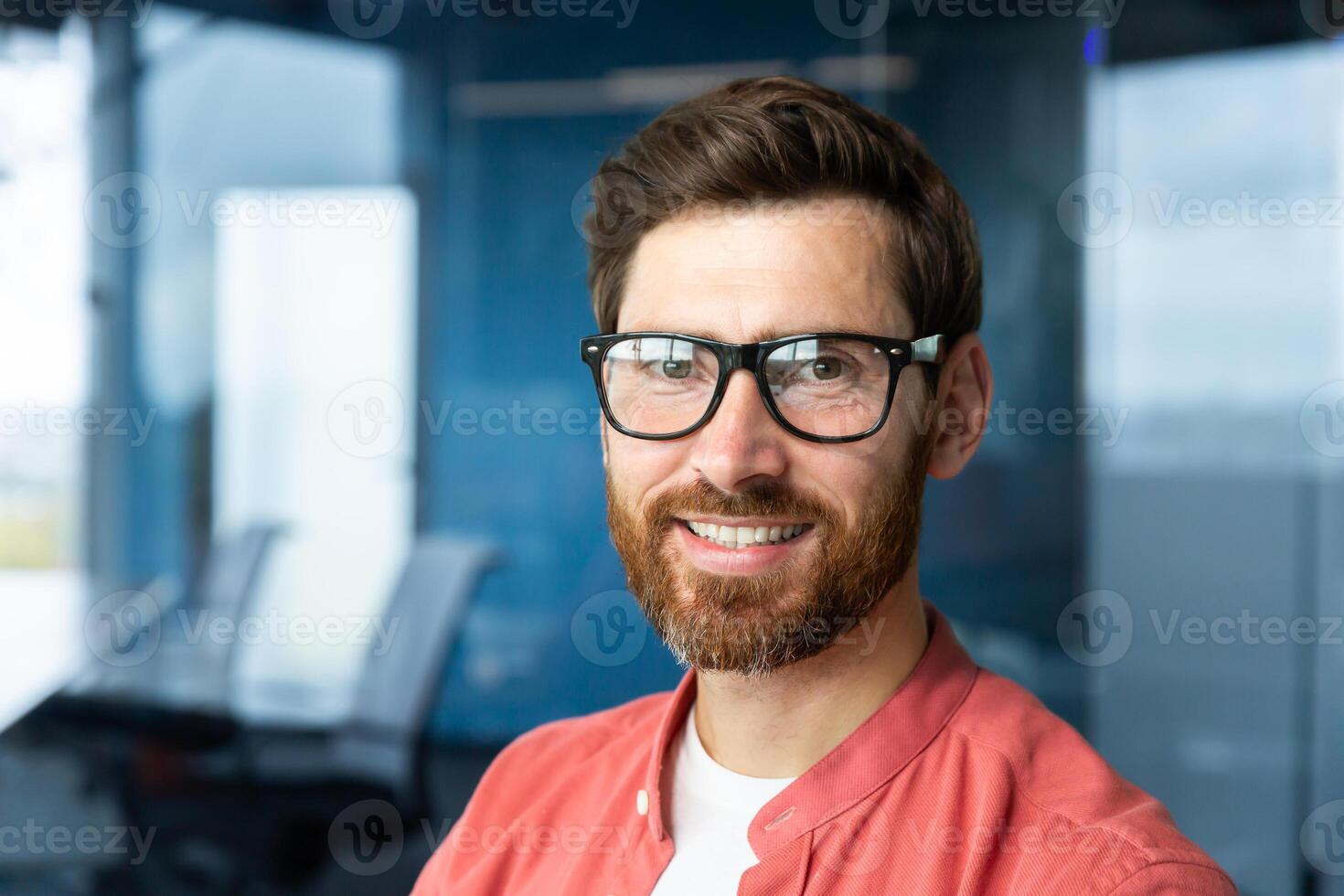 detailopname portret van jong zakenman in rood shirt, volwassen Mens in bril glimlachen en op zoek Bij camera binnen kantoor in de buurt venster, geslaagd investeerder gelukkig met prestaties en mooi zo investering. foto