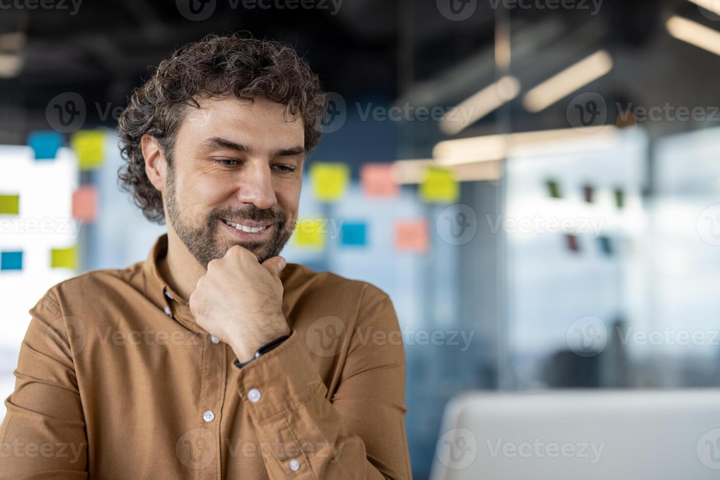 professioneel mannetje in een gewoontjes overhemd werken en glimlachen terwijl op zoek Bij de laptop in een helder kantoor milieu met kleverig aantekeningen in de achtergrond. foto