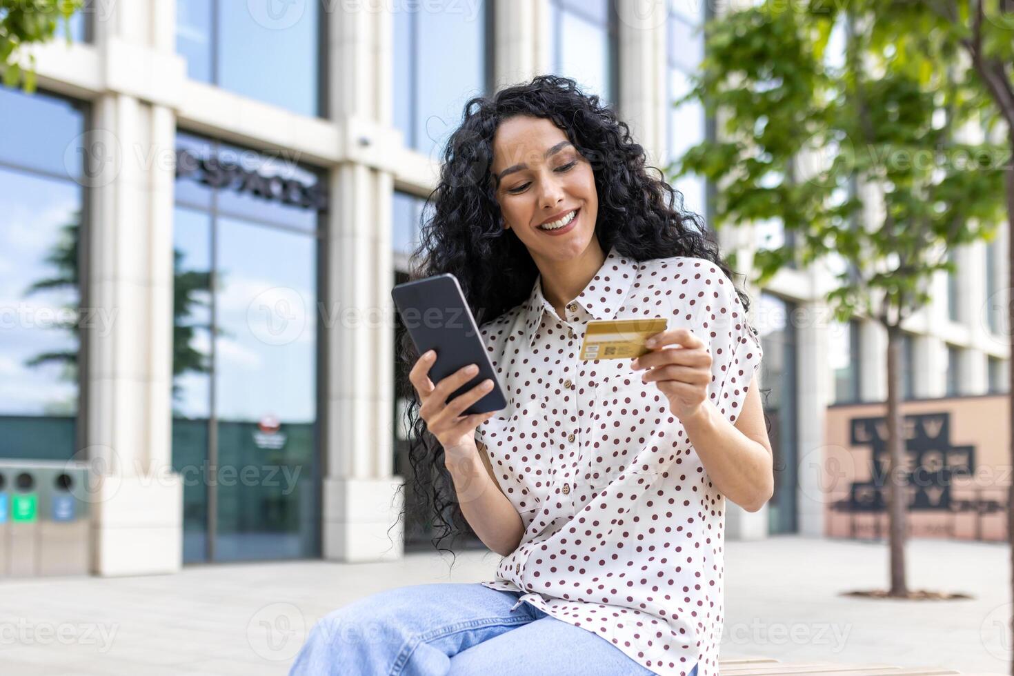 jong mooi vrouw wandelen in de stad, tevreden Latijns Amerikaans vrouw met gekruld haar- houdt telefoon en bank credit kaart in handen, maakt online aankopen gelukkig boeken Diensten en kiest online. foto