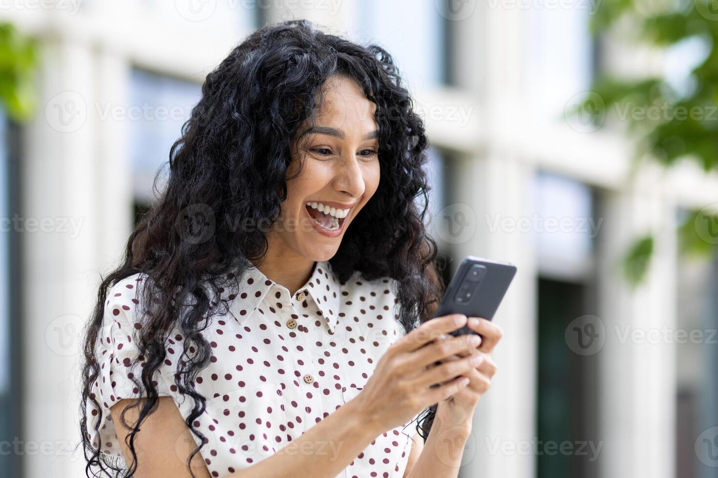 blij vrouw met gekruld haar- lachend terwijl op zoek Bij smartphone, buitenshuis met wazig achtergrond, overbrengen geluk en digitaal levensstijl. foto