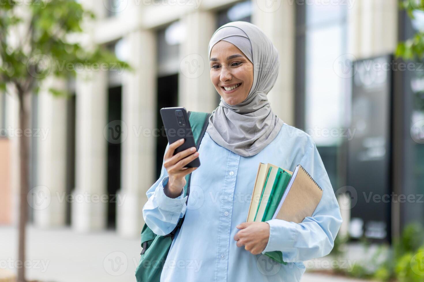 jong moslim vrouw in hijab wandelen buiten Universiteit campus, vrouw leerling glimlachen tevreden gebruik makend van app Aan telefoon, rugzak Aan terug en boeken in handen. foto