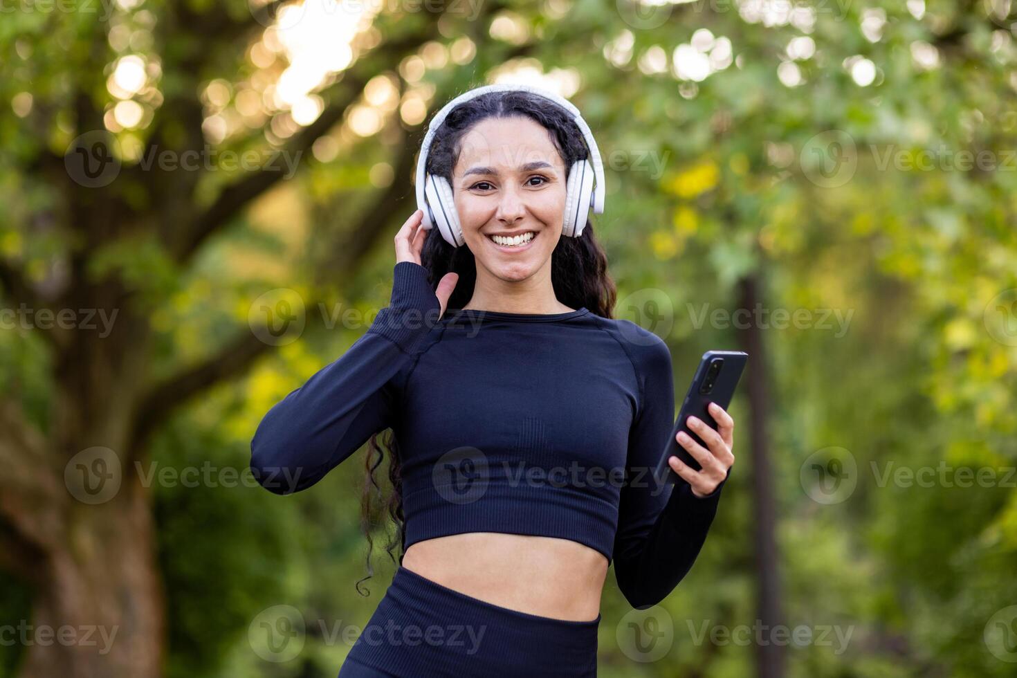 portret van vrolijk vrouw atleet in strak passend sportkleding luisteren naar muziek- in oortelefoons verbonden naar mobiel telefoon. gekruld dame beoefenen gezond levensstijl en genieten van oefenen Aan Open lucht. foto