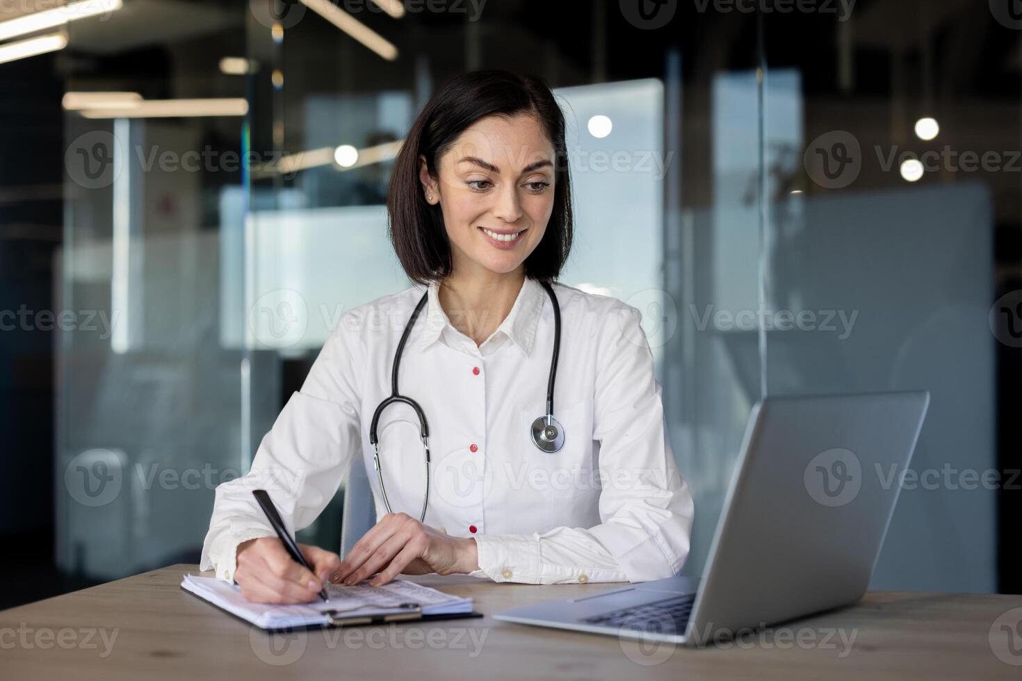 aantrekkelijk vrouw therapeut in professioneel uniform op zoek Bij laptop scherm en maken aantekeningen door hand- Aan klembord. hedendaags ziekenhuis arbeider het verstrekken van online Diensten en consultaties voor cliënt. foto