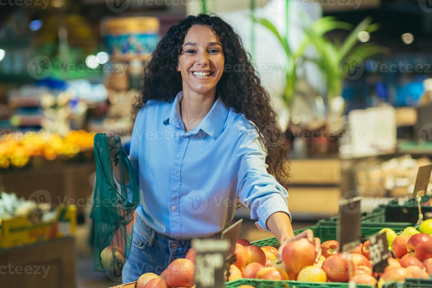 portret van gelukkig vrouw klant in supermarkt, spaans vrouw kiest appels en fruit glimlachen en op zoek Bij camera, met kruidenier mand kiest goederen foto