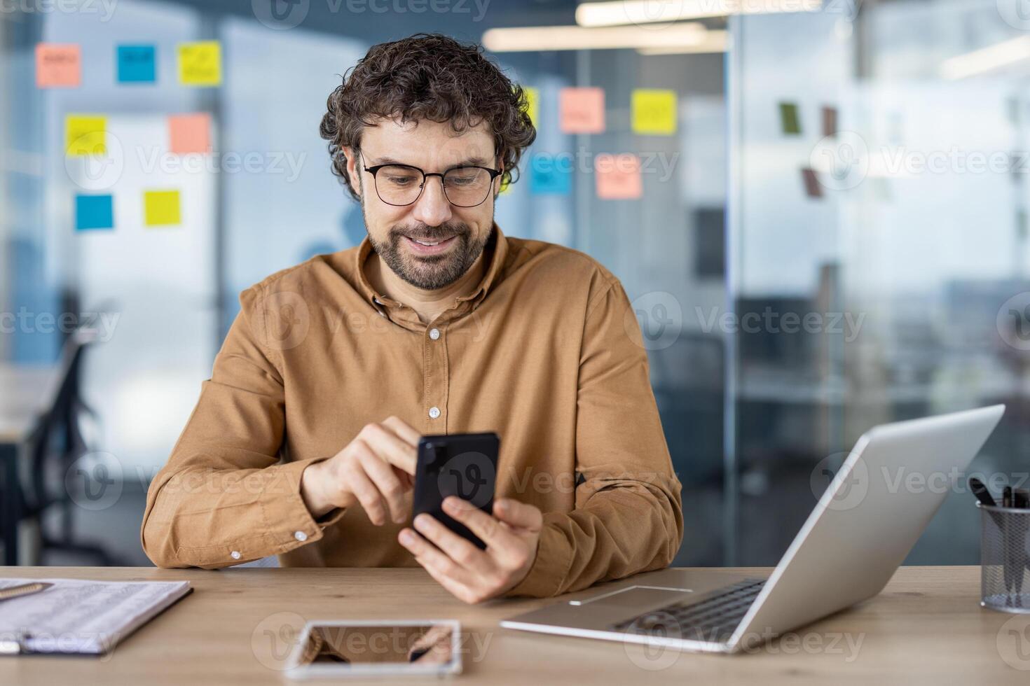 gefocust mannetje professioneel in een gewoontjes overhemd gebruik makend van smartphone en laptop Bij een modern werkplaats met kleverig aantekeningen in de achtergrond. foto