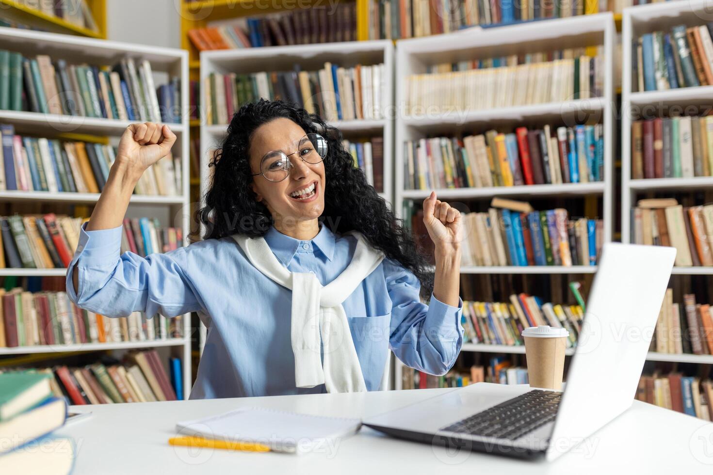 opgewonden jong vrouw in een bibliotheek verhogen haar vuisten in zege, met laptop en koffie kop Aan de tafel. een symbool van academisch succes en geluk. foto