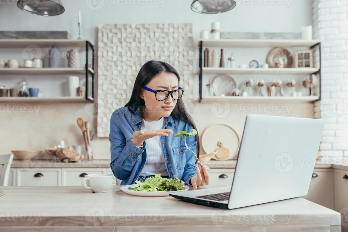 ontevreden Aziatisch vrouw met online diëten, vrouw zittend in keuken Bij huis aan het eten salade voor gewicht verlies, gebruik makend van laptop naar visie aanbevelingen en leren over eetpatroon en gezond aan het eten. foto