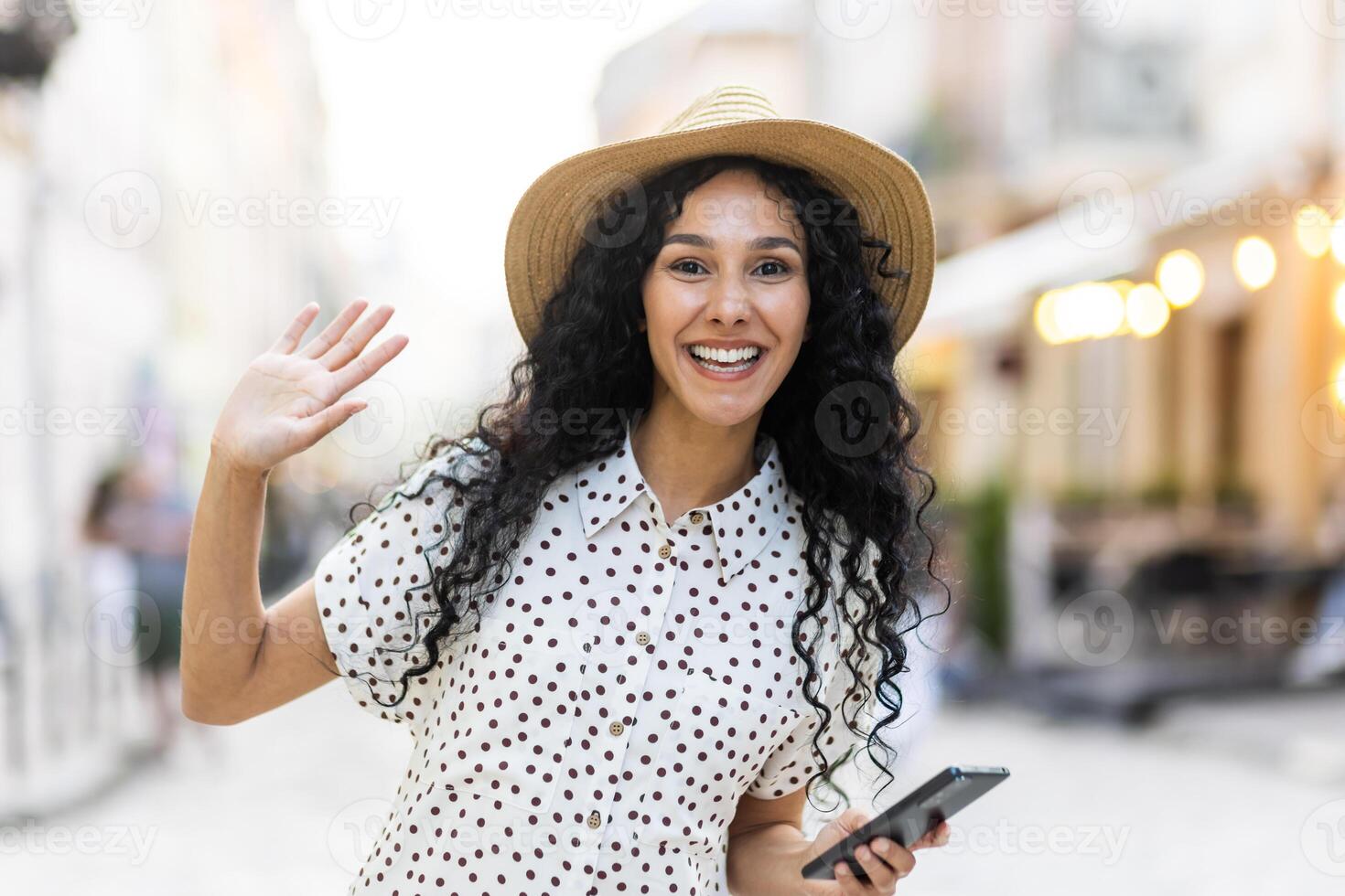 portret van een gelukkig jong Indisch vrouw wandelen in de stad Aan een zonnig dag, Holding de telefoon in haar hand, glimlachen en groet, golvend Bij de camera. foto