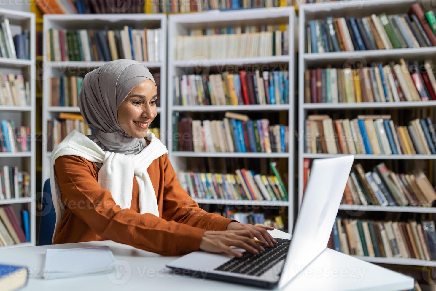 jong mooi leerling in hijab aan het studeren online van een afstand in Universiteit bibliotheek, vrouw typen Aan laptop toetsenbord zoeken voor informatie en voorbereidingen treffen voor examen. foto