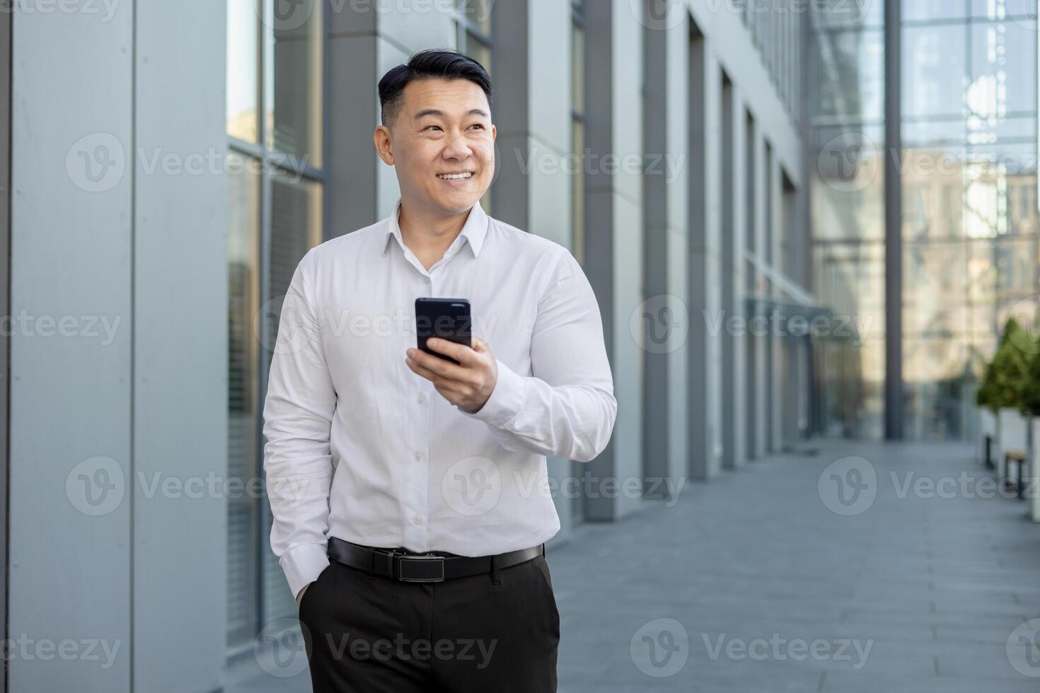 glimlachen jong Aziatisch Mens wandelen in de buurt kantoor centrum buiten, Holding telefoon in hand- en op zoek weg. foto