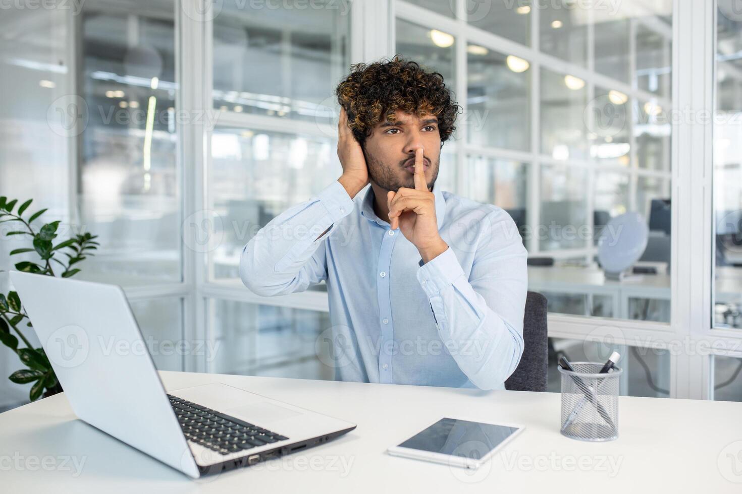 jong professioneel Mens in een blauw overhemd zittend Bij een wit kantoor bureau, maken een stilte gebaar met een vinger naar lippen, omringd door modern kantoor omgeving. foto
