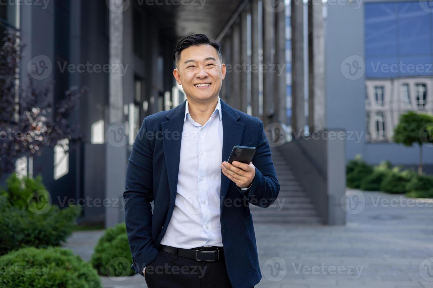 portret van Aziatisch geslaagd jong Aziatisch Mens zakenman, advocaat, politicus in pak staand in de buurt kantoor gebouw Holding telefoon en hand- in zak, glimlachen op zoek Bij camera. foto