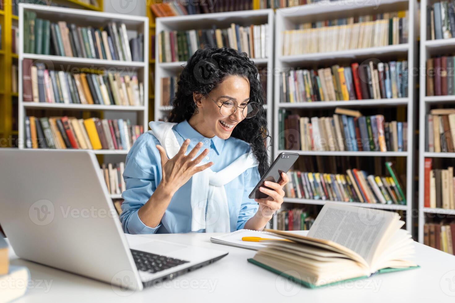 blij spaans leerling met bril uitdrukken geluk terwijl lezing haar smartphone, omringd door boeken in de bibliotheek. foto