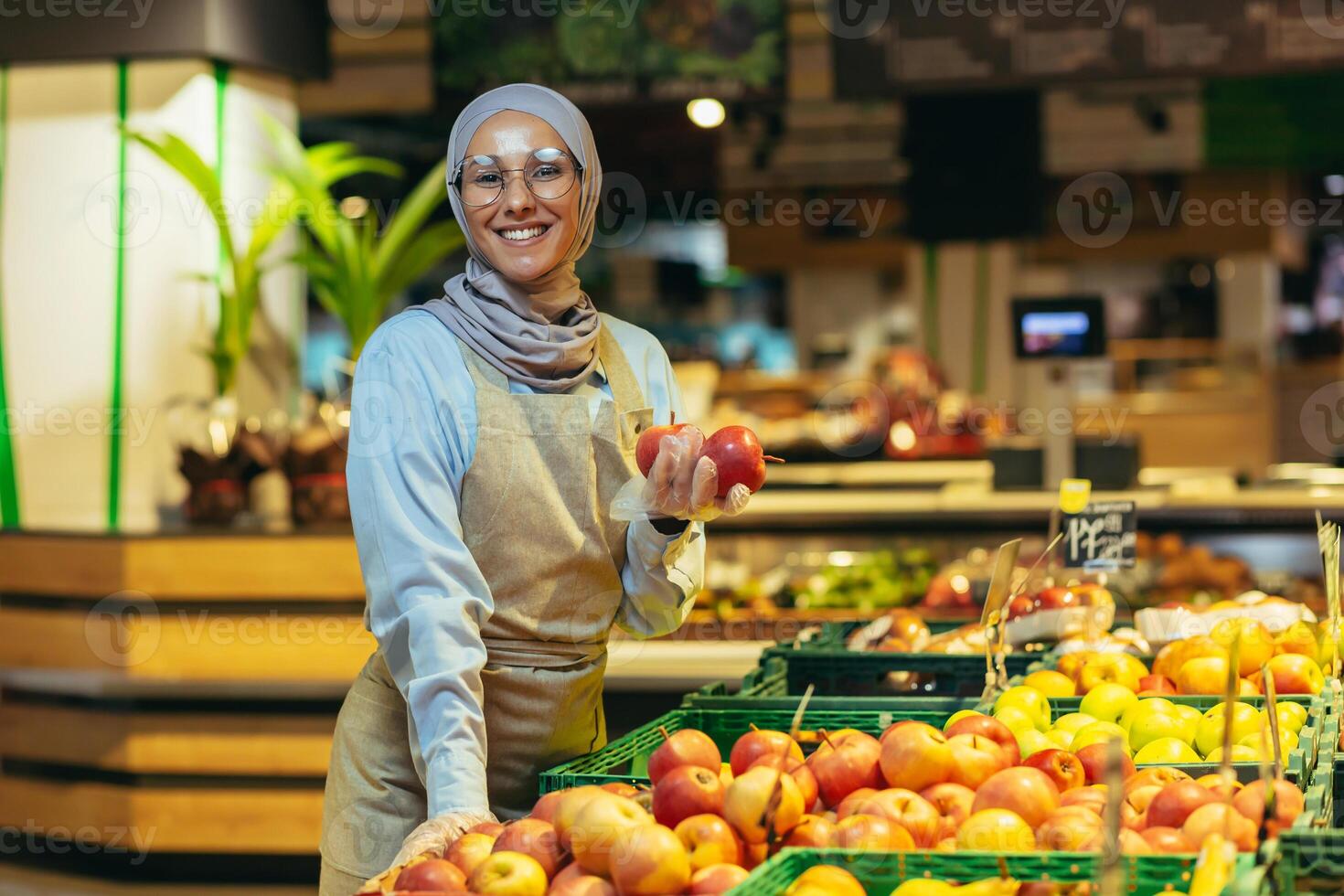 portret van verkoopster in supermarkt, gelukkig vrouw in hijab glimlachen en op zoek Bij camera, verkoper Holding appels in groente sectie, moslim vrouw in bril en schort tussen schappen met goederen foto