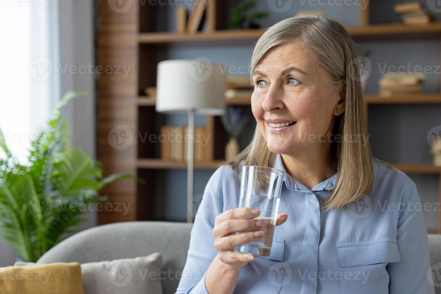 een volwassen vrouw met grijs haar- glimlacht hartelijk terwijl Holding een glas van water, gezeten comfortabel in een goed verlicht leven kamer versierd met planten en boeken. foto