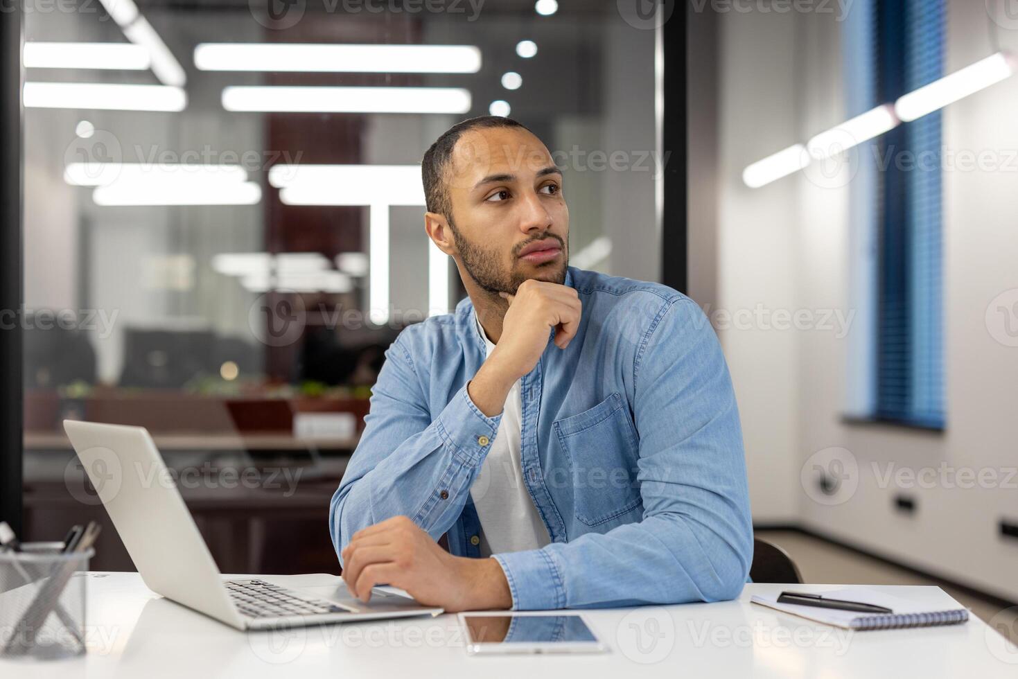 een nadenkend mannetje ondernemer in een denim overhemd zit Bij zijn laptop in een hedendaags kantoor instelling, staren door de venster, reflecterend Aan bedrijf strategieën of overweegt nieuw ideeën. foto