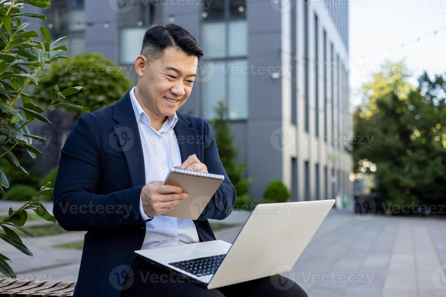 vrolijk Aziatisch mannetje professioneel nemen aantekeningen en gebruik makend van laptop terwijl werken buiten kantoor terrein. foto