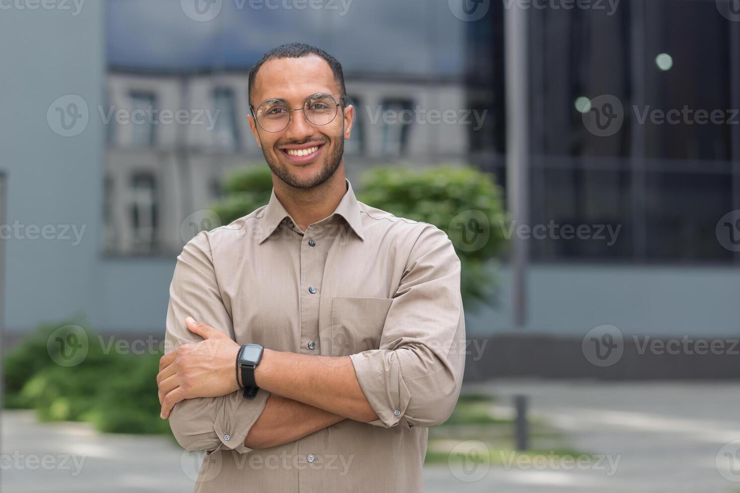 portret van jong spaans leerling Mens glimlachen en op zoek Bij camera in de buurt Universiteit campus met armen gekruiste vervelend bril en overhemd foto