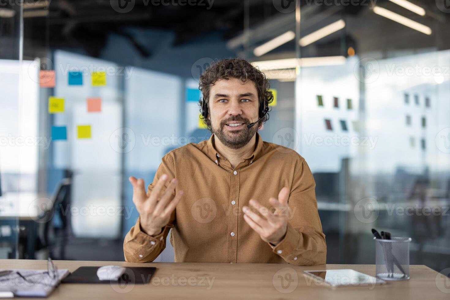 glimlachen mannetje professioneel met koptelefoon innemend in een bedrijf gesprek in een hedendaags werkruimte met kleverig aantekeningen in achtergrond. foto