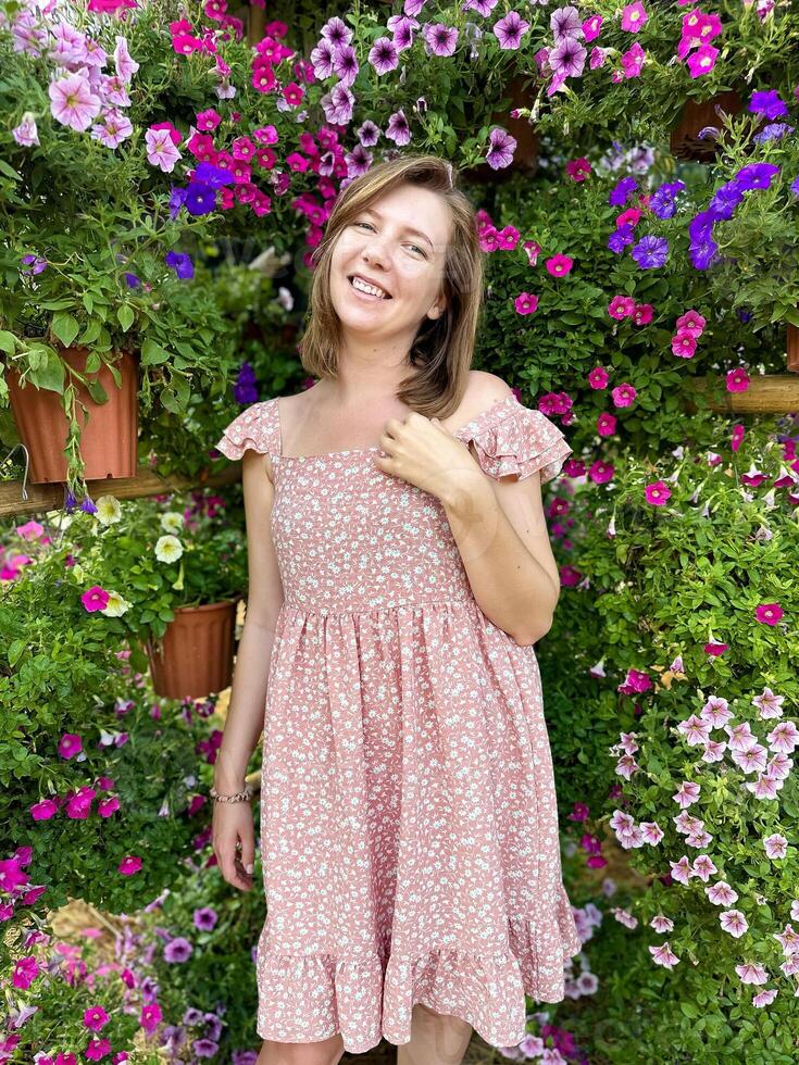 charmant jong vrouw glimlachen in een bloeiend tuin, omringd door levendig roze en Purper bloemen, vervelend een gewoontjes roze bloemen jurk foto