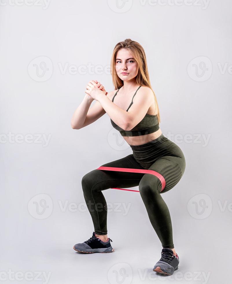 Sportieve vrouw doet squats met behulp van rubberen weerstandsband geïsoleerd op een grijze achtergrond foto