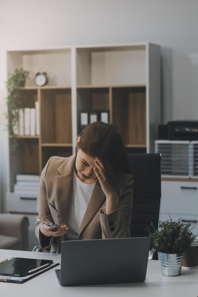 Aziatisch Dames zittend in een kantoor met spanning en oog spanning moe, portret van verdrietig ongelukkig moe gefrustreerd teleurgesteld dame lijden van migraine zittend Bij de tafel, ziek arbeider concept foto