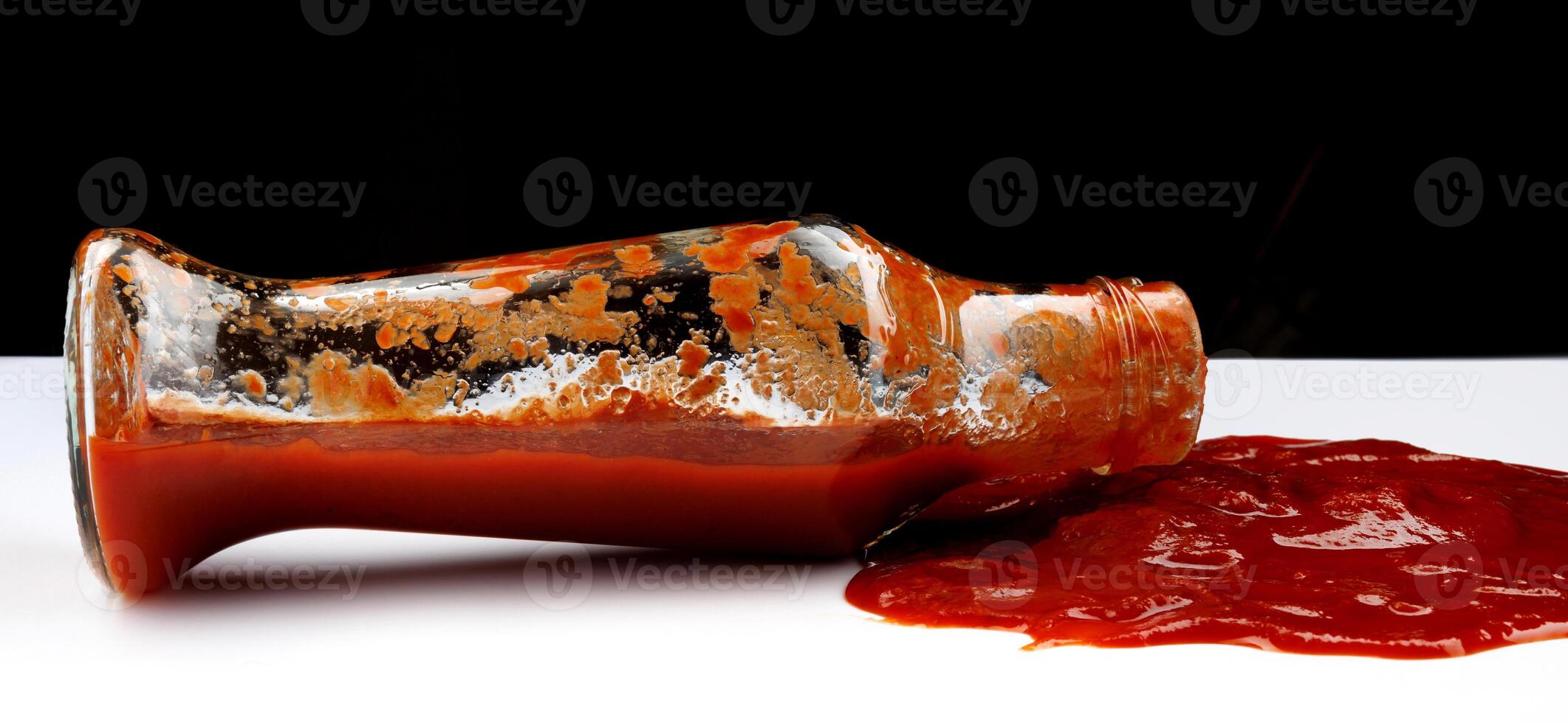 tomaat saus . een glas fles met ketchup en een plas van ketchup gemorst in de omgeving van het. foto