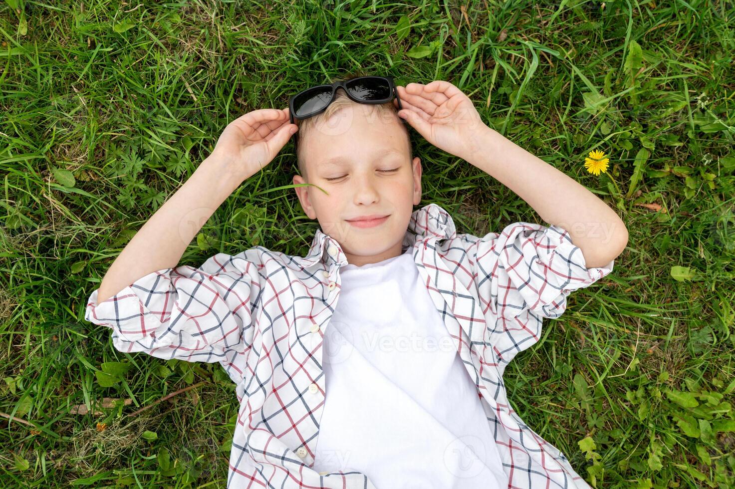 een schattig jongen leugens in de gras met zonnebril en een overhemd met zijn ogen Gesloten foto