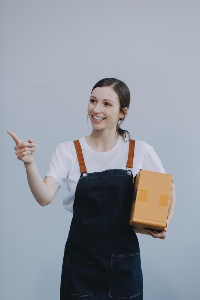 glimlachen Aziatisch vrouw in gewoontjes kleren Holding een karton doos mockup terwijl staand tegen een geïsoleerd wit achtergrond. Verzending bedrijf concept foto