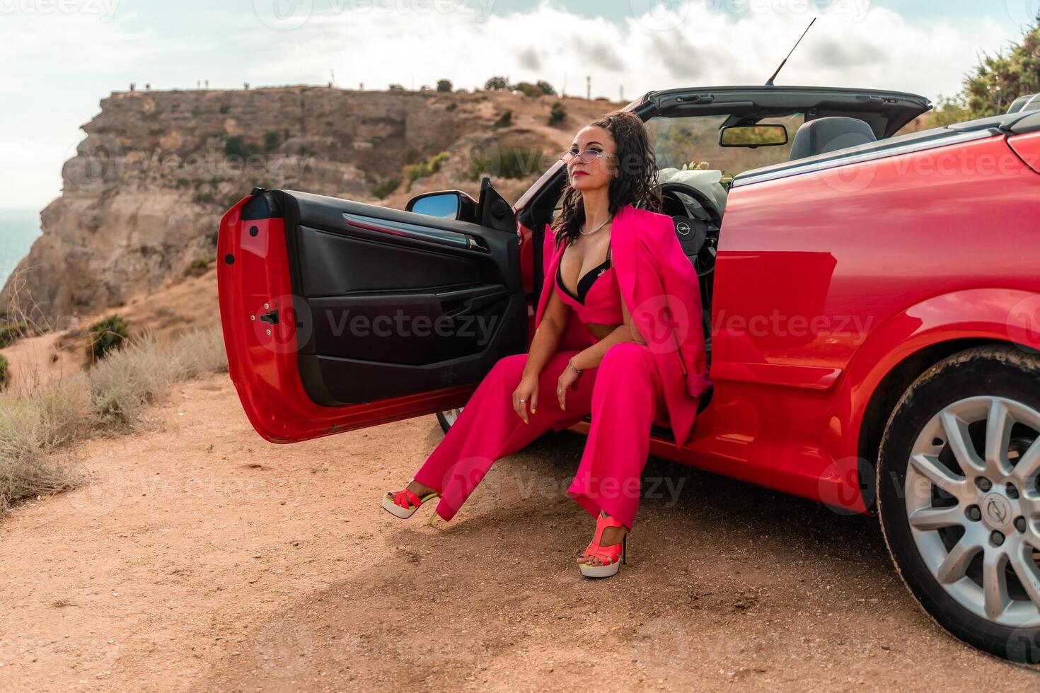 een vrouw in een roze pak zit in de Open deur van een rood converteerbaar. de tafereel is reeks Aan een rotsachtig heuvel met uitzicht de oceaan. de vrouw is poseren voor een foto. foto