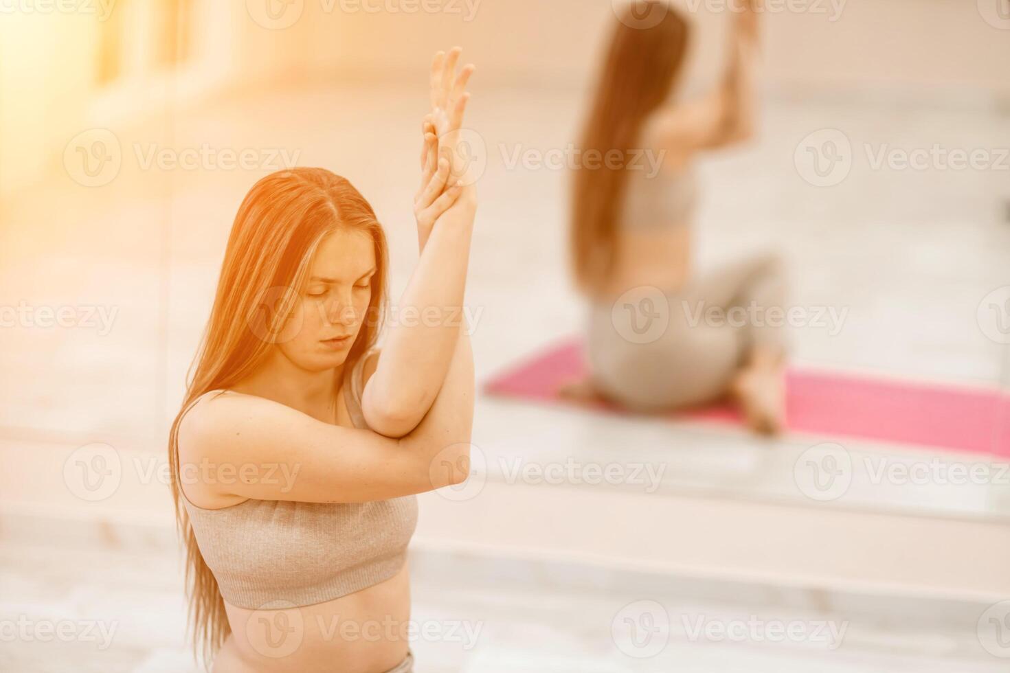 jong vrouw aan het doen yoga in de Sportschool. een meisje met lang haar- en in een beige trainingspak staat in een koe houding Aan een roze tapijt. een vrouw presteert gomukhasana. foto