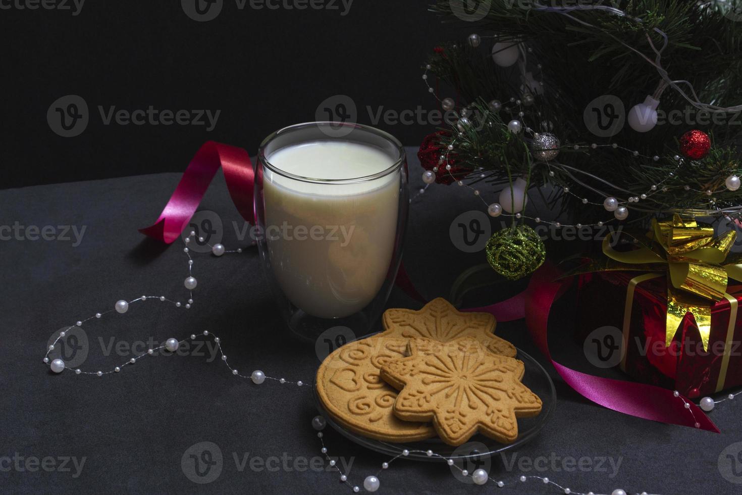 peperkoek en melk voor de kerstman. kerstcompositie met peperkoekkoekjes en melk op een zwarte achtergrond met een kerstboom en een cadeau. foto