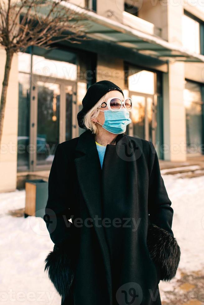 senior stijlvolle vrouw in baret en elegante zwarte jas en met medisch masker buiten wandelen. lockdown, pandemie, beschermingsconcept foto