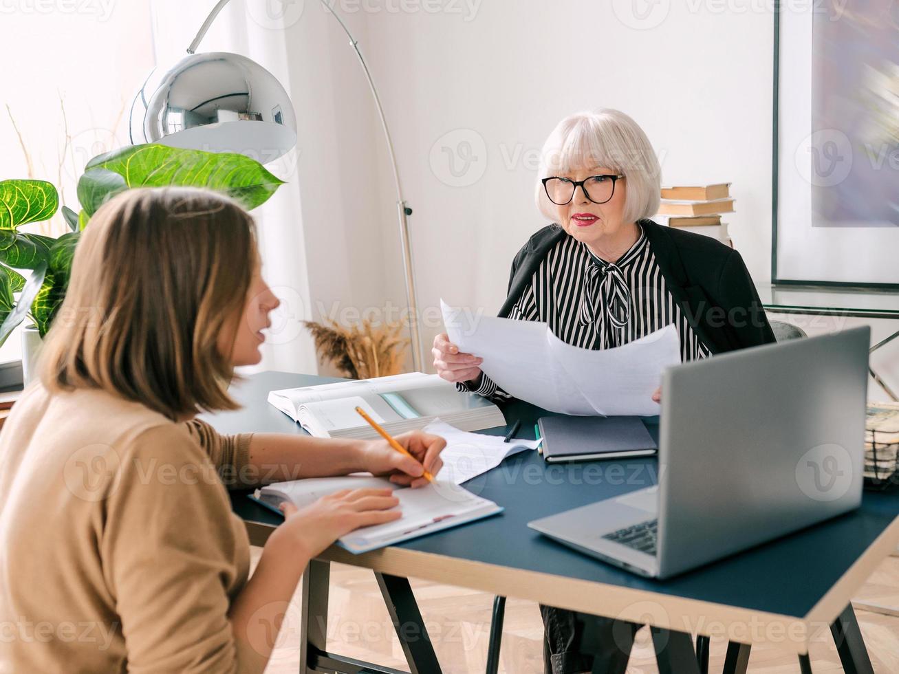 senior stijlvolle vrouw met jonge vrouw die werktaken op kantoor bespreekt. bedrijf, communicatie, werk, leeftijden, samenwerking, mentorconcept foto