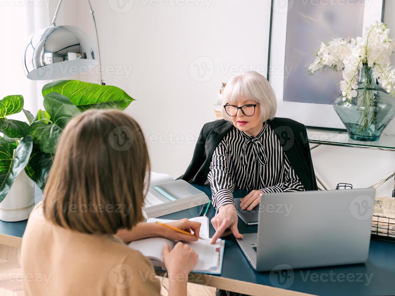 senior stijlvolle vrouw met jonge vrouw die werktaken op kantoor bespreekt. bedrijf, communicatie, werk, leeftijden, samenwerking, mentorconcept foto