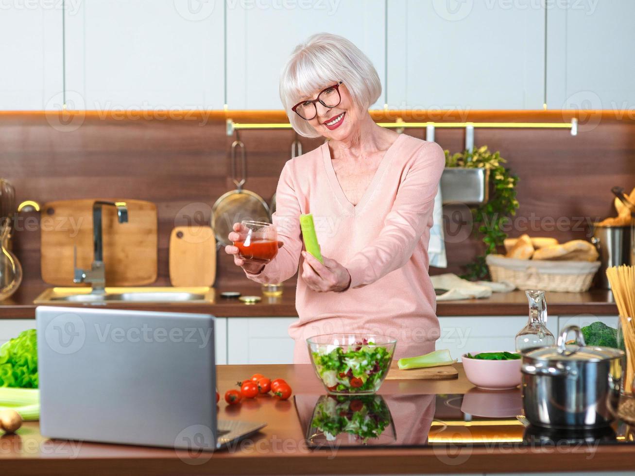 senior vrolijke vrouwenvideoblogger die selderij en tomatensap toont in haar keuken. rauw, vegetarisch, dieet, blogger, voedingsdeskundige keukenconcept foto