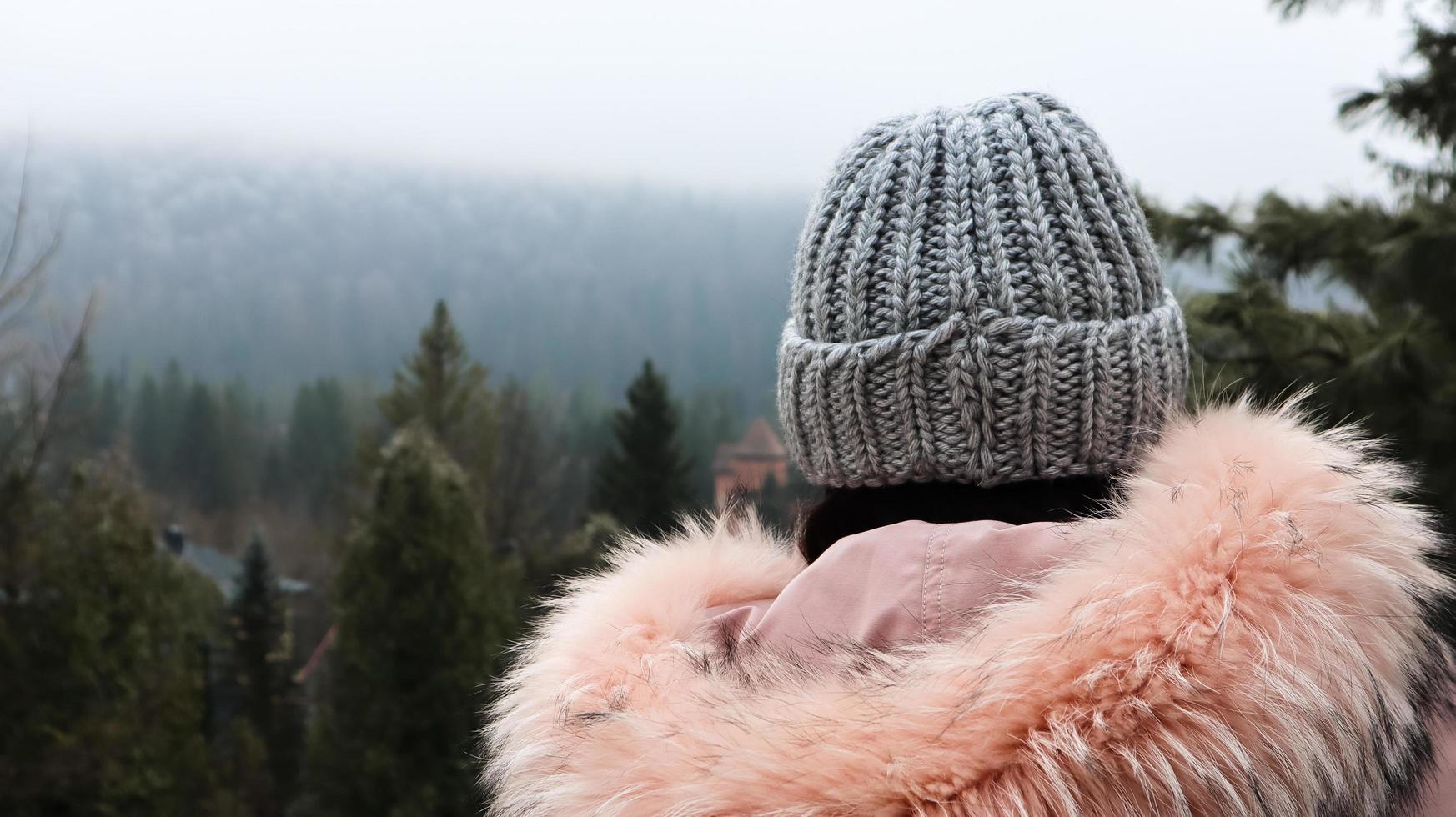 achteraanzicht van een vrouw bergtoerist in een roze jas met bont en een grijze gebreide muts tegen een achtergrond van bergen op zoek naar het herfstlandschap van het bos en de bewolkte hemel. foto