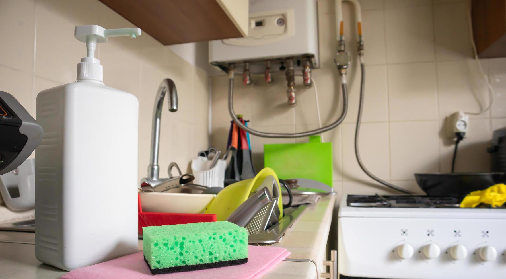 groene spons en vloeibare zeepdispenser voor het afwassen op een vuile gootsteen, volledig met servies en keukengerei. afwassen in de keuken met de hand met afwasmiddel en een spons. foto