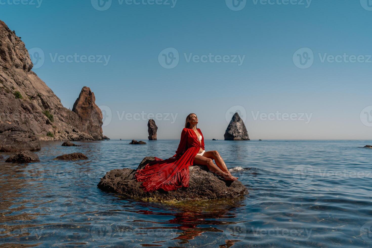 mooi sensueel vrouw in een vliegend rood jurk en lang haar, zittend Aan een rots bovenstaand de mooi zee in een groot baai. foto